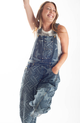 A woman wearing a pair of oversized blue denim overall-style jumpsuit with a tonal geometric pattern, posing against a solid white background.