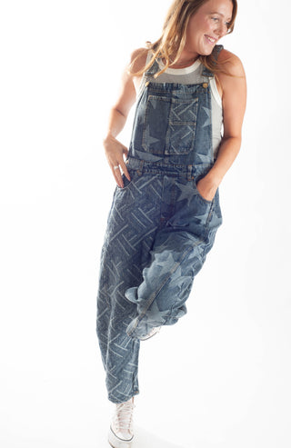 A woman modeling a pair of blue denim overall-style pants with a faded geometric pattern, worn over a white tank top and white sneakers against a plain white background.