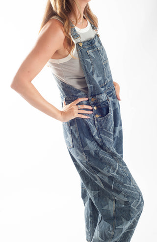 A model wearing blue denim overall-style pants with a tonal geometric star pattern, styled over a white tank top against a white studio background.