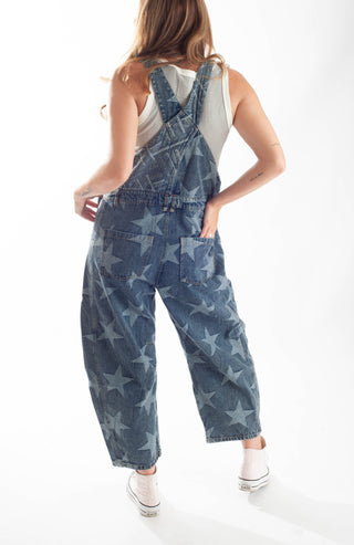A model from behind wearing blue denim overall with a light blue star pattern, styled with a white tank top and white sneakers against a white studio background.