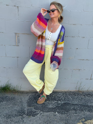 Woman wearing a colorful cardigan and yellow pants leaning against a gray wall.