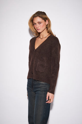 A woman modeling a dark brown fuzzy knit cardigan with a V-neck and front pockets, paired with dark blue jeans, against a plain light gray background.