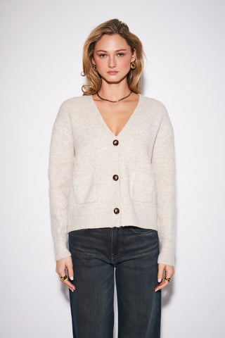 A woman modeling a light beige knit cardigan with dark brown buttons, paired with dark wash jeans, against a plain light gray studio background.