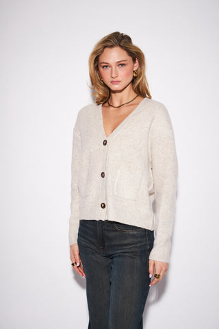 A woman modeling a light beige knit cardigan with dark buttons, paired with dark blue jeans, against a plain white studio background.