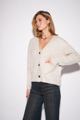 A woman modeling a light beige knit cardigan with dark buttons, paired with dark blue jeans, against a plain white studio background.