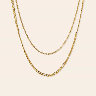 Two gold-tone chain necklaces layered on a light cream background. The top necklace is a delicate cable chain, while the bottom necklace is a slightly thicker figaro link chain.