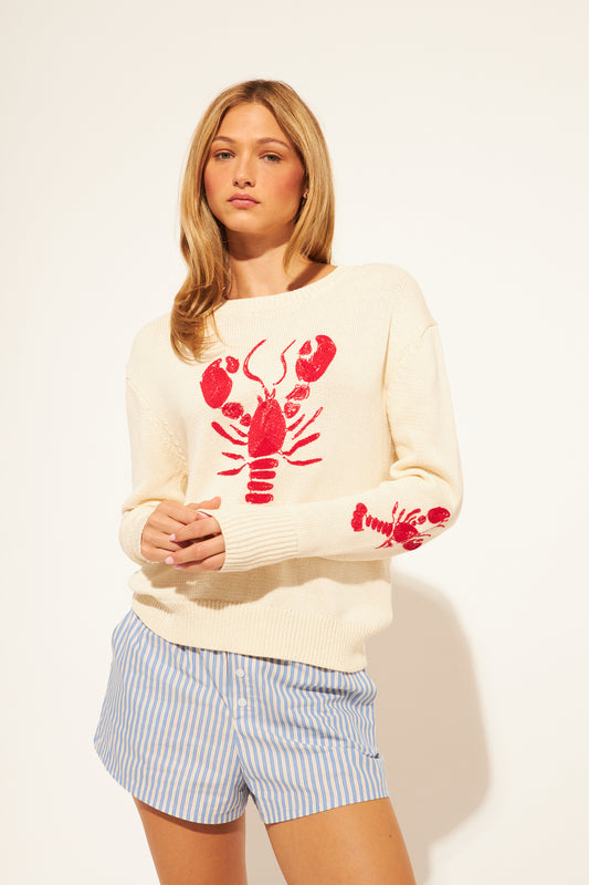 Woman wearing a cream sweater with a red lobster design and blue shorts on a white background