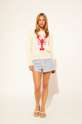 Woman wearing a cream sweater with a red lobster design and checkered shorts on a white background