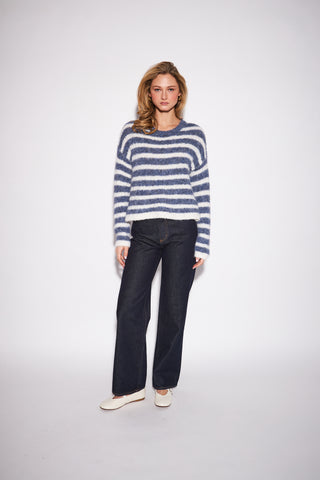 A woman modeling a fuzzy, long-sleeve knit sweater with horizontal navy and off-white stripes, paired with dark wide-leg jeans and white flat shoes, against a plain light gray studio background.