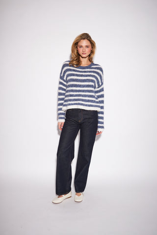 A woman modeling a fuzzy blue and white striped long-sleeve sweater, paired with dark wide-leg jeans and white slip-on shoes, against a plain light gray studio background.