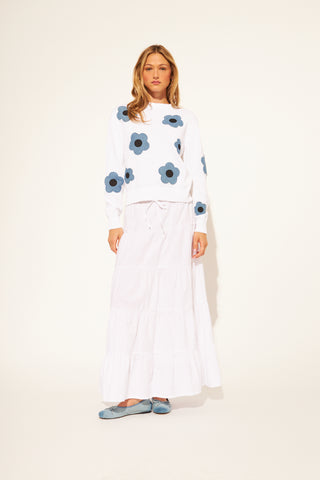 Woman wearing a white sweater with blue floral patterns and white pants on a light beige background