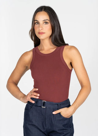A woman modeling a sleeveless crew-neck tank top in a deep burgundy color, paired with dark blue jeans, against a plain light gray studio background.