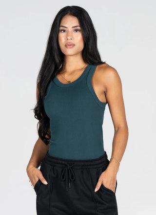 A woman modeling a dark teal green ribbed tank top paired with black sweatpants against a light gray studio background.