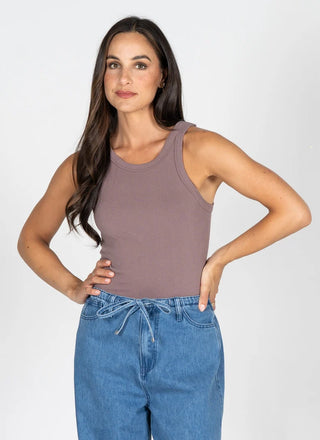A woman modeling a sleeveless scoop-neck tank top in a muted mauve-brown color, paired with blue jeans, against a plain light gray studio background.