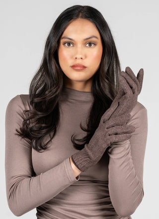 A woman with long dark hair wearing a taupe long-sleeve top and matching taupe knit gloves, posing against a light gray studio background.