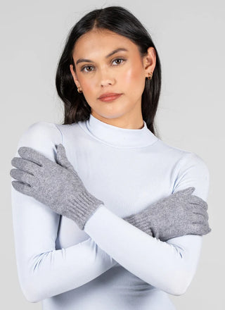 A woman wearing gray knit gloves and a light blue mock-neck long-sleeve top, posing with her arms crossed against a solid light gray background.