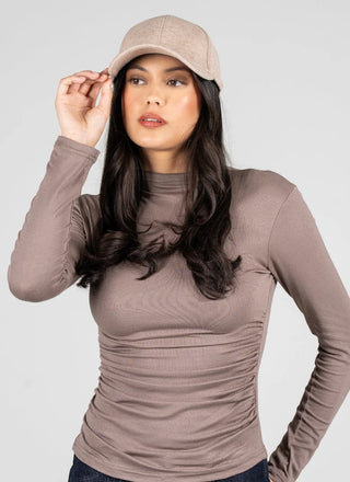 A woman wearing a beige baseball cap and a fitted, long-sleeve ruched top in a taupe-brown color, posed against a solid light gray background.