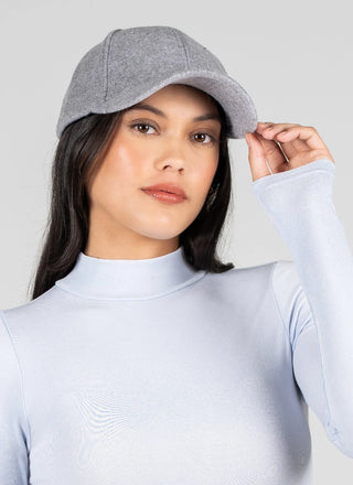 A woman wearing a light gray wool cap and a pale blue mock-neck long-sleeve top, posing against a solid light gray background.
