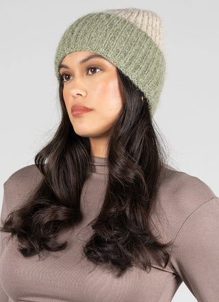 A woman wearing a ribbed knit beanie hat in sage green and cream, paired with a taupe ribbed top, photographed against a solid light gray background.