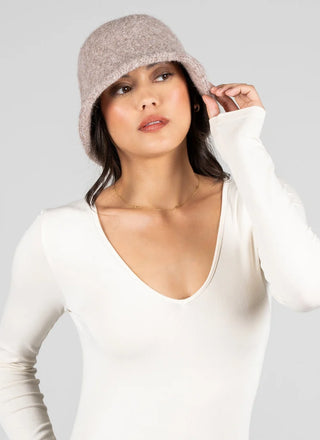 A woman wearing a light taupe knit bucket hat and a white long-sleeve V-neck top, posing against a solid light gray background.