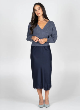 A woman modeling a two-piece outfit consisting of a heathered slate blue long-sleeve knit top and a dark navy midi skirt with a tassel fringe hem, paired with light gray heels, against a plain light gray studio background.
