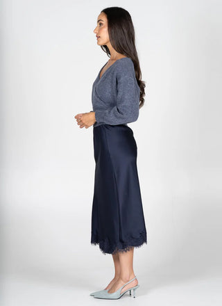 A woman stands in profile wearing a gray wrap-style knit top and a dark navy midi skirt with lace trim, paired with pale blue slingback heels, against a light gray studio background.