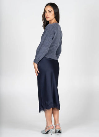 A woman modeling a gray knit sweater and a navy midi skirt with lace trim, standing against a light gray studio background.