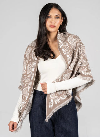 A woman modeling a taupe and white paisley patterned shawl with fringed edges, worn over a white long-sleeve top and dark blue jeans against a solid gray background.