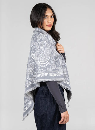 A woman wearing a gray and white paisley patterned shawl over a dark gray long-sleeve top and dark blue jeans, photographed against a solid light gray studio background.