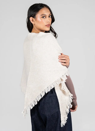 A woman wearing a cream-colored fringed poncho wrap over a brown top and dark blue jeans, photographed against a solid light gray background.