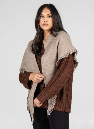 A woman modeling a brown and beige knit poncho-style cardigan with fringed edges, worn over a brown cable-knit sweater against a light gray studio background.