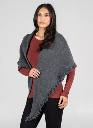 A woman modeling a dark gray fringed shawl or poncho over a rust-colored long-sleeve top and black pants, photographed against a plain light gray studio background.