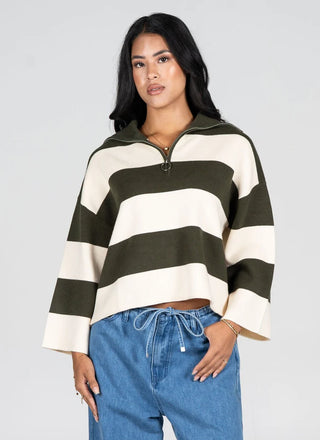 A woman modeling a cropped, long-sleeve half-zip pullover sweater with wide horizontal stripes in dark olive green and cream, paired with blue jeans against a light gray studio background.