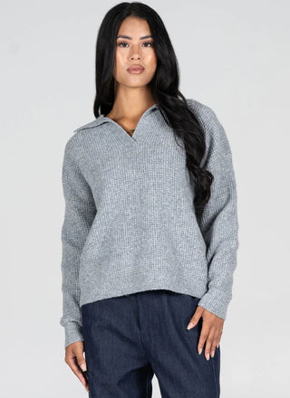 A woman modeling a light gray ribbed knit polo sweater with a split collar, paired with dark blue pants, against a plain light gray studio background.