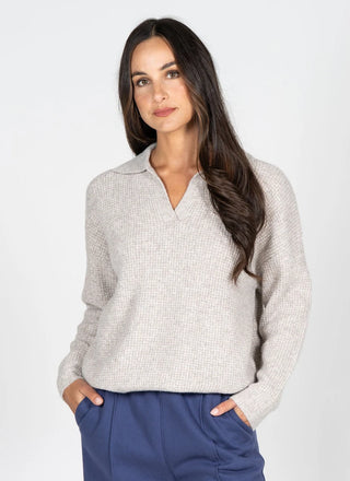 A woman modeling a light beige textured knit polo sweater with a V-neck, paired with navy blue pants, against a plain light gray background.