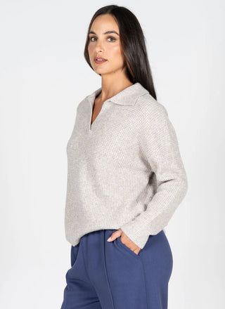 A woman modeling a light gray textured knit polo sweater with a collar and split neckline, paired with navy blue trousers, against a plain white studio background.