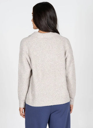 A woman viewed from the back wearing a light beige textured knit sweater and navy blue trousers, set against a plain light gray studio background.