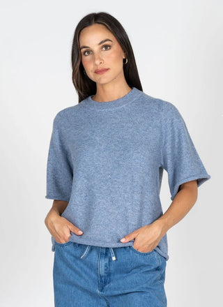A woman wearing a heathered blue short-sleeve knit sweater and blue jeans, standing against a light gray studio background.