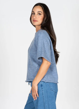 A woman wearing a heathered light blue short-sleeve knit top and blue jeans, shown from a side-back angle against a plain white studio background.