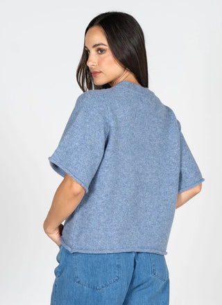 A woman shown from the back wearing a heathered blue short-sleeve knit top and blue jeans against a plain light gray studio background.