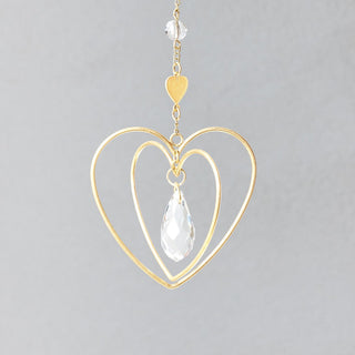 A gold-colored heart-shaped suncatcher with a clear teardrop-shaped crystal hanging in the center, attached by a chain to a metal hook.