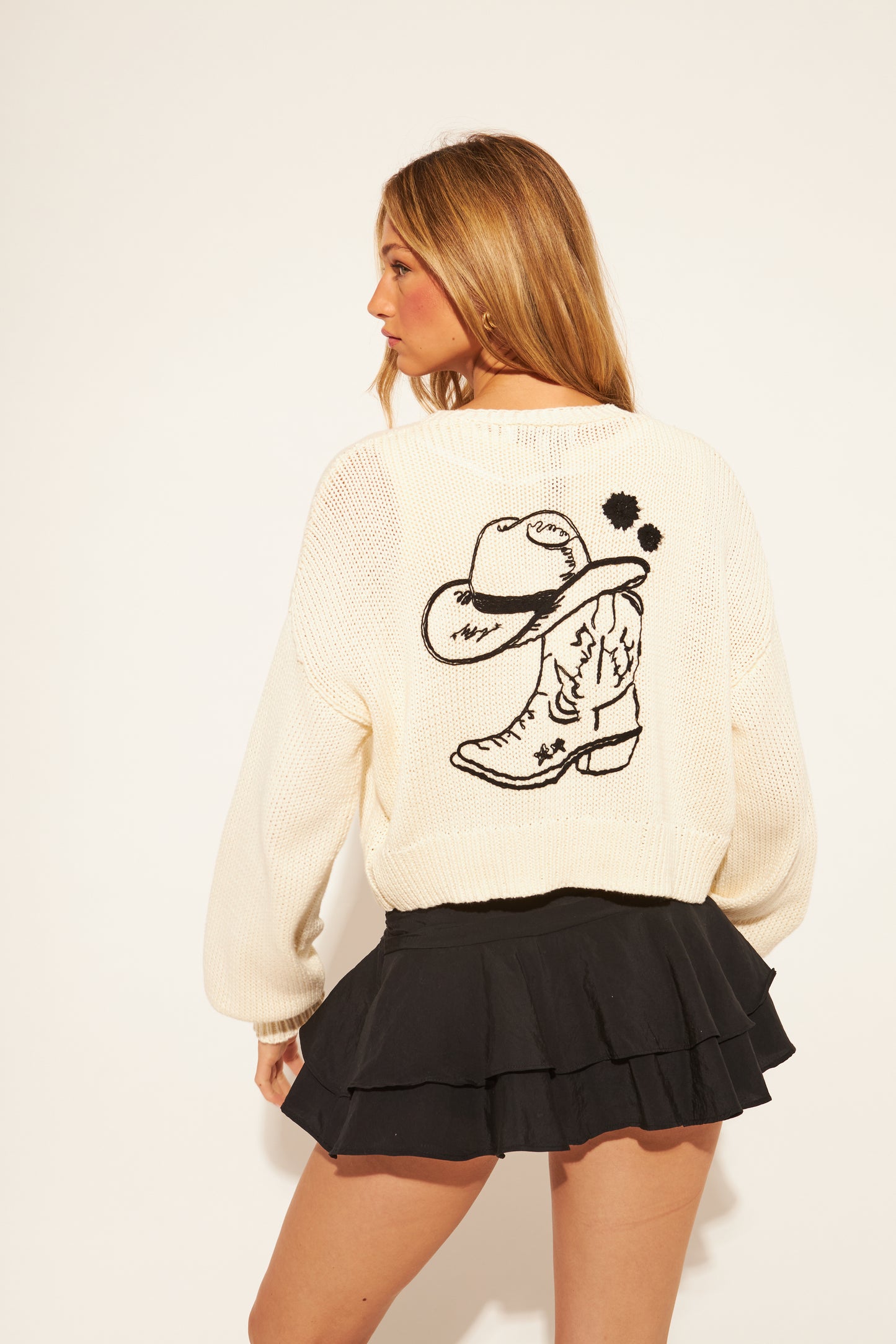 Woman wearing a cream sweater with a black boot design on the back, standing against a plain background.