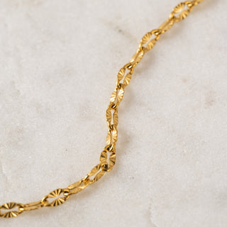 A gold-colored necklace with a textured chain design, featuring small sunburst patterns, laid out on a marble surface.