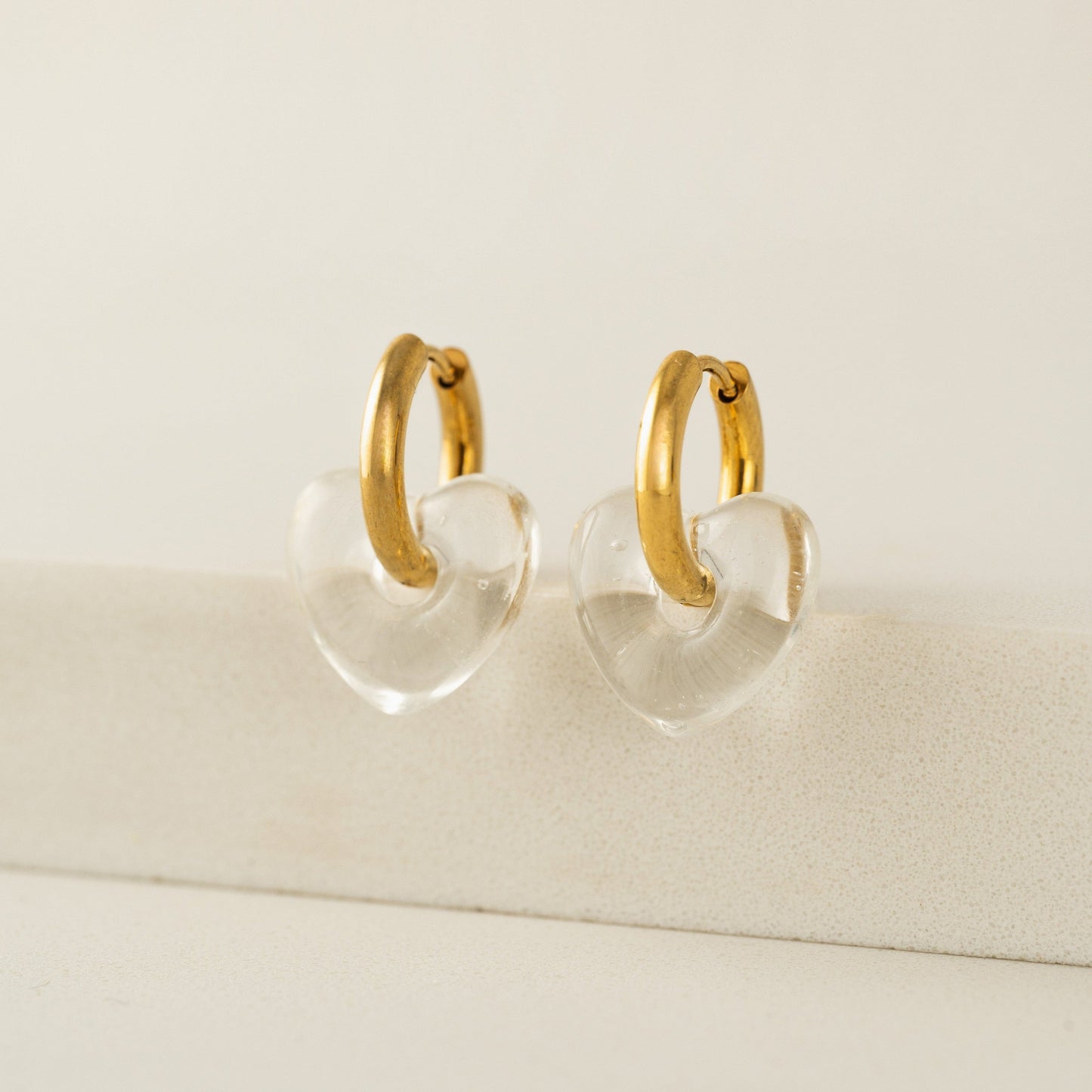 Gold hoop earrings with clear heart-shaped centers on a beige background