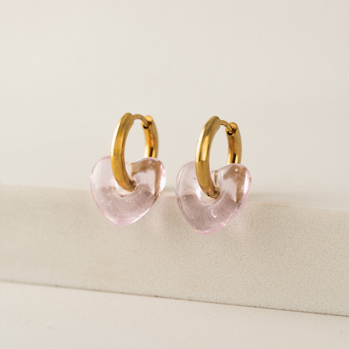 Gold hoop earrings with pink heart-shaped charms on a beige surface