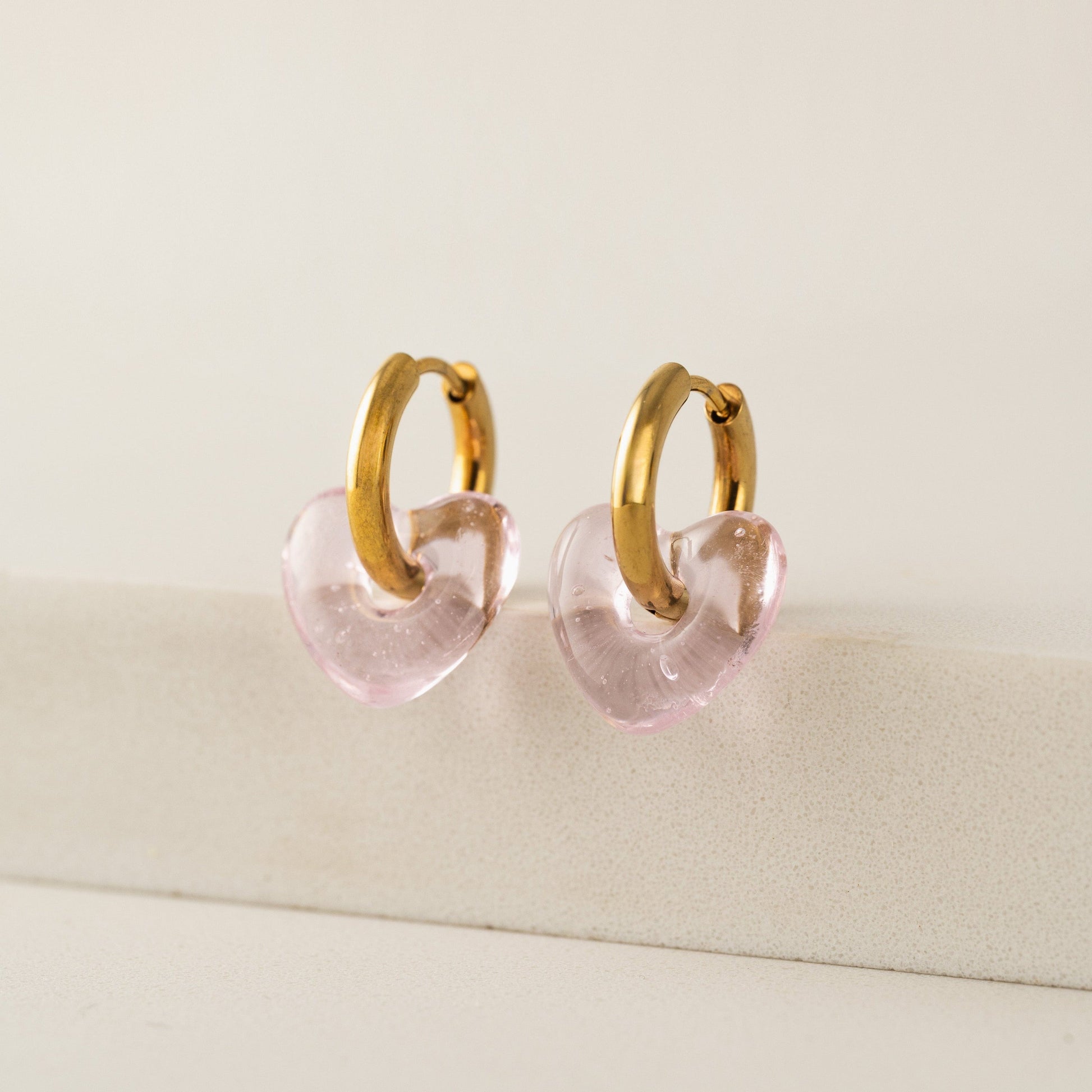 Gold hoop earrings with pink heart-shaped charms on a beige surface