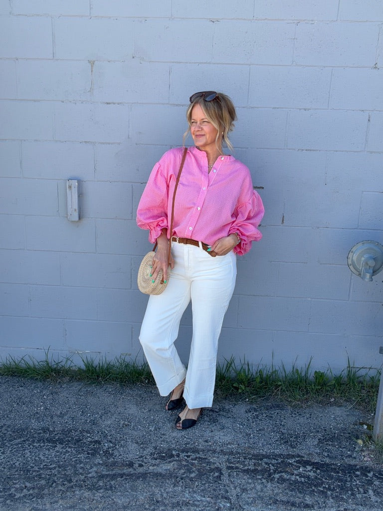 Woman in a pink blouse and white pants standing against a light blue wall.
