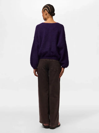 A model shown from the back wearing a fuzzy, oversized deep purple sweater and dark brown wide-leg trousers, photographed against a clean white studio background.