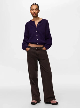 A model stands against a light gray studio background wearing a dark purple cropped button-front cardigan, dark brown wide-leg trousers, and pink slip-on shoes.