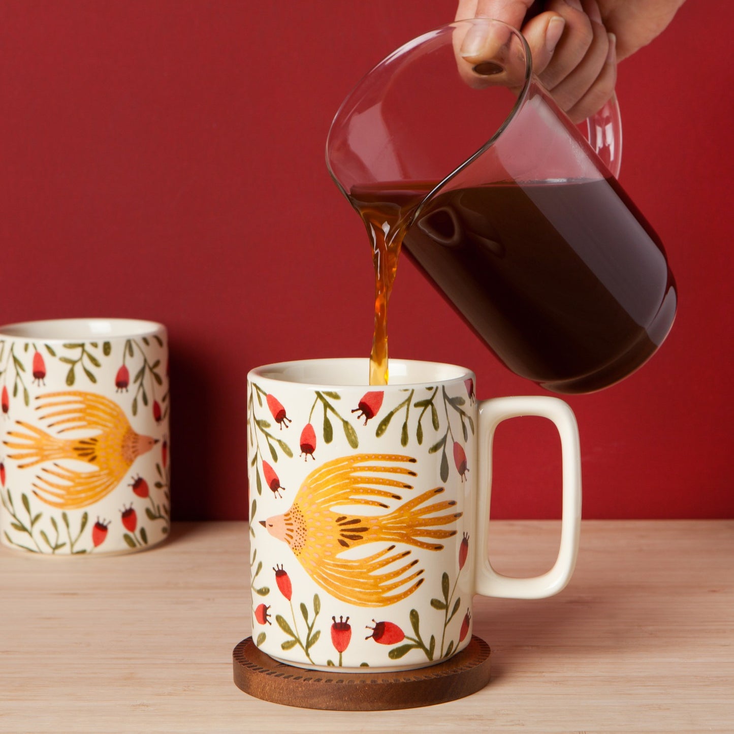 Person pouring coffee into a mug with bird design on a wooden table against a red background